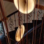 Looking down through tan cords at two lit alabaster discs of this Clérachic pendant, over a spiral stair with scrolled iron balusters.