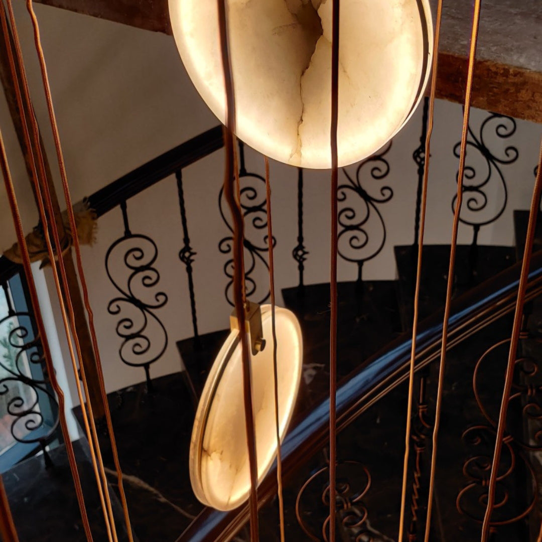 Looking down through tan cords at two lit alabaster discs of this Clérachic pendant, over a spiral stair with scrolled iron balusters.