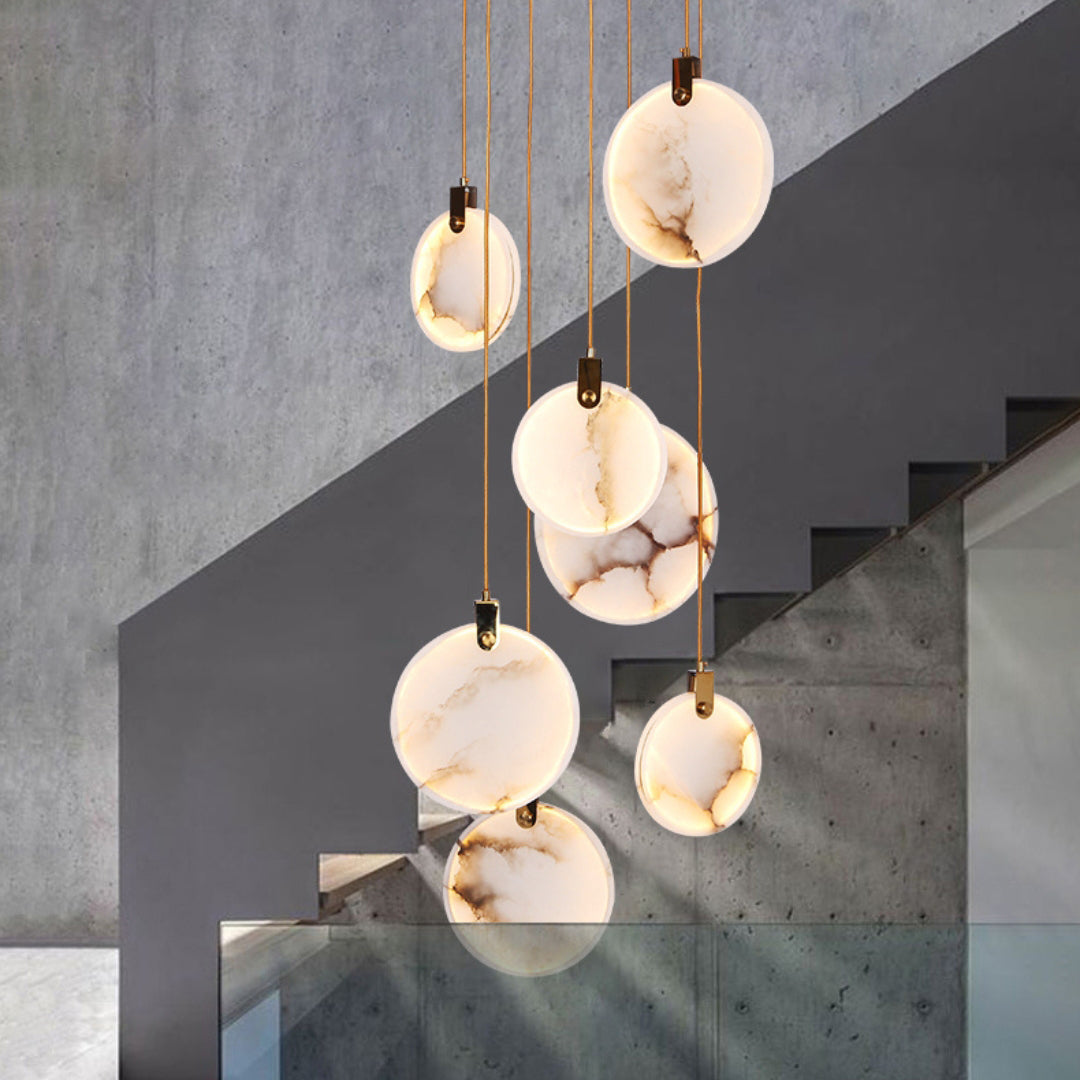 Clérachic alabaster discs hang at different heights in front of a grey concrete wall and a dark staircase with glass panels.