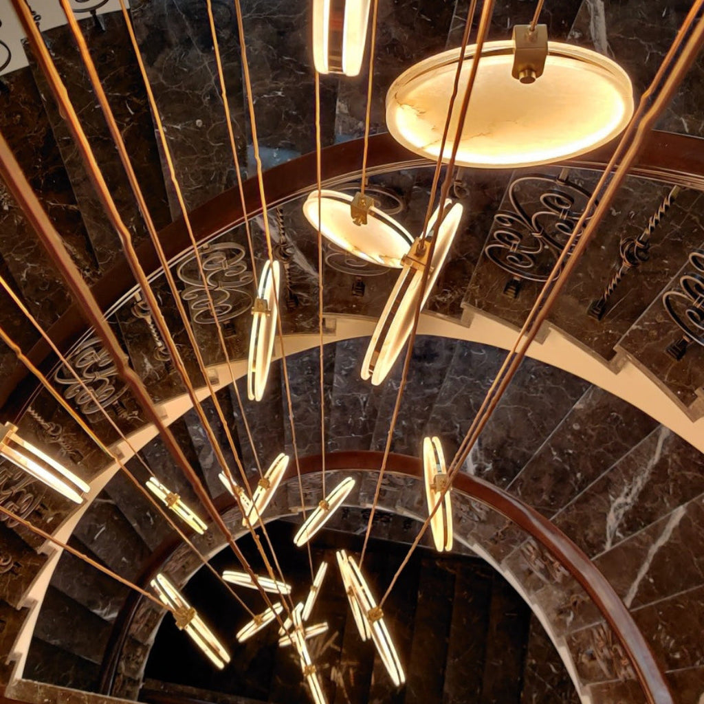 Looking up a spiral staircase of dark marble, edge-on Clérachic alabaster discs glow among long tan cords.