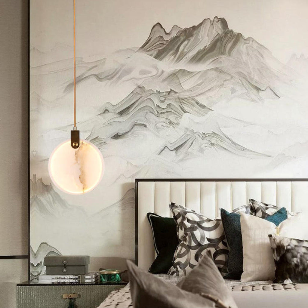 One Clérachic alabaster disc pendant on a tan cord hangs before a mountain landscape mural, above a bed with patterned cushions.