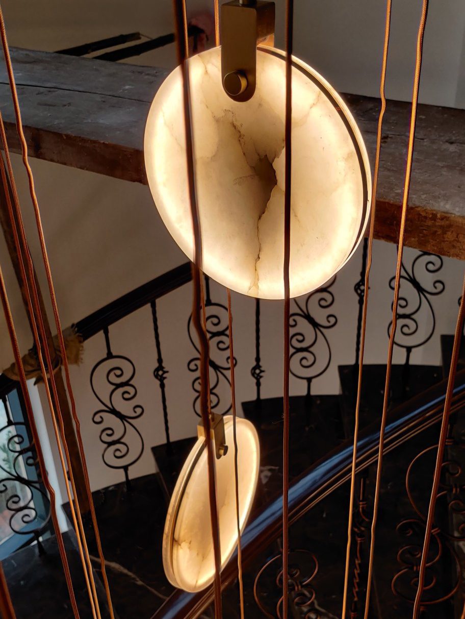 Close view from below of a lit alabaster disc and its brass hanger on this Clérachic pendant, above a stair with scrolled iron railings.