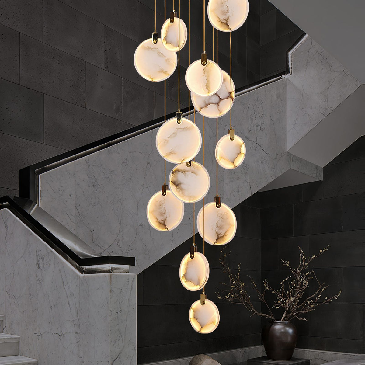 Lit Clérachic alabaster discs hang beside a stair of grey marble with a recessed handrail light, with a vase of branches below.