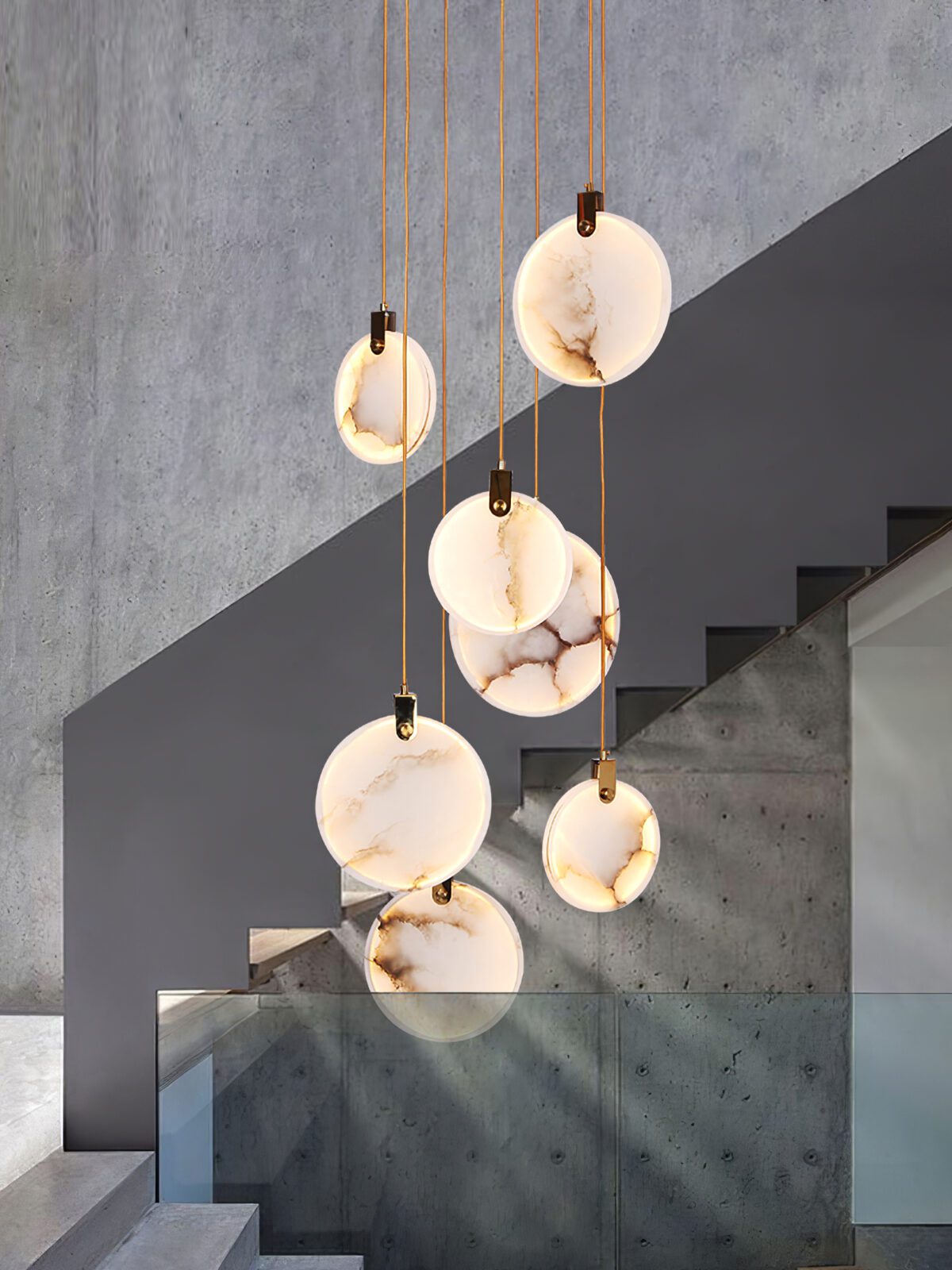 Lit alabaster discs of this Clérachic pendant hang in front of a concrete stair wall and a glass balustrade.