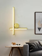 Gold Clérachic wall light with a vertical and a horizontal tube and a round disc, lit on a pale wall above a grey sofa.