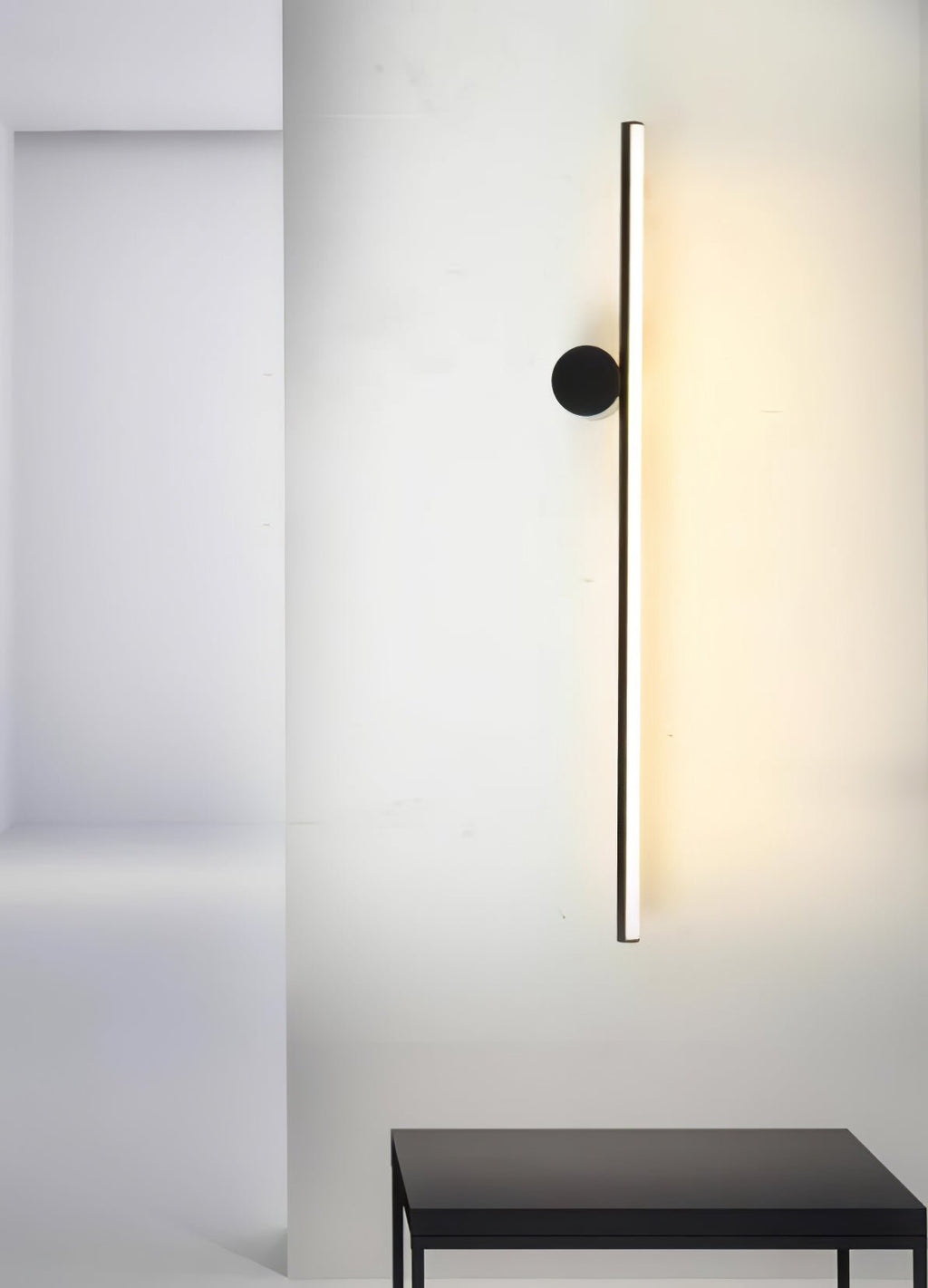 Black vertical tube of this Clérachic wall light with a round black disc, glowing on a white wall beside a black table.