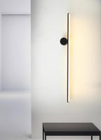 Black vertical tube of this Clérachic wall light with a round black disc, glowing on a white wall beside a black table.