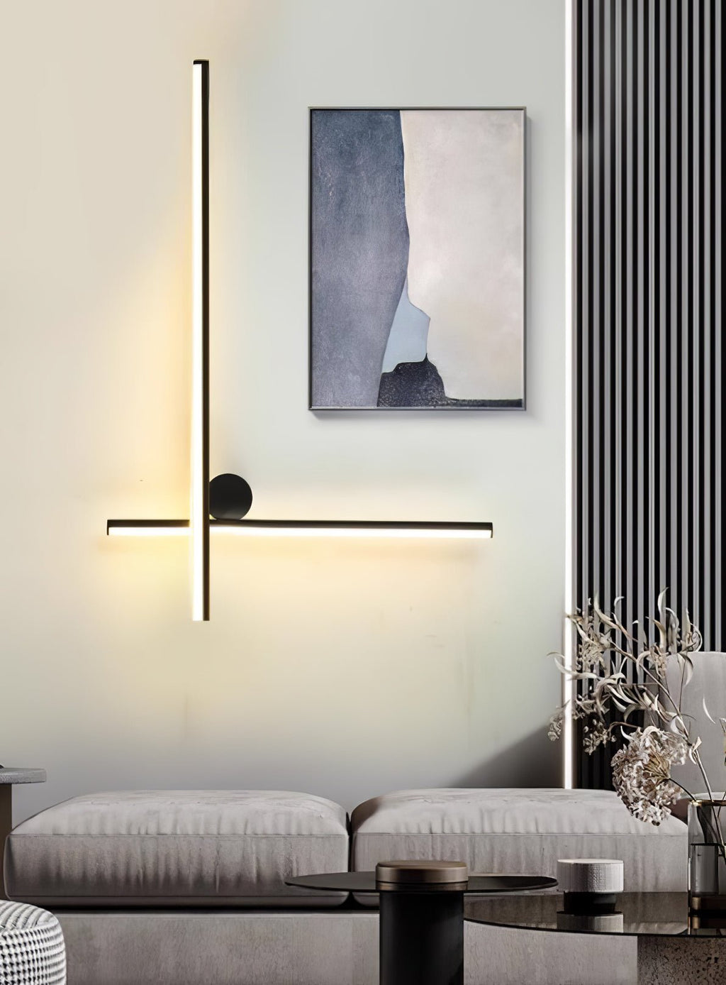Black Clérachic wall light with a vertical and a horizontal tube and a disc, lit above a grey sofa with a framed print beside it.