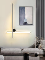 Black Clérachic wall light with a vertical and a horizontal tube and a disc, lit above a grey sofa with a framed print beside it.