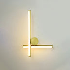 Gold Clérachic wall light with a vertical and a horizontal tube crossing, a gold disc and a warm glow on a pale wall.