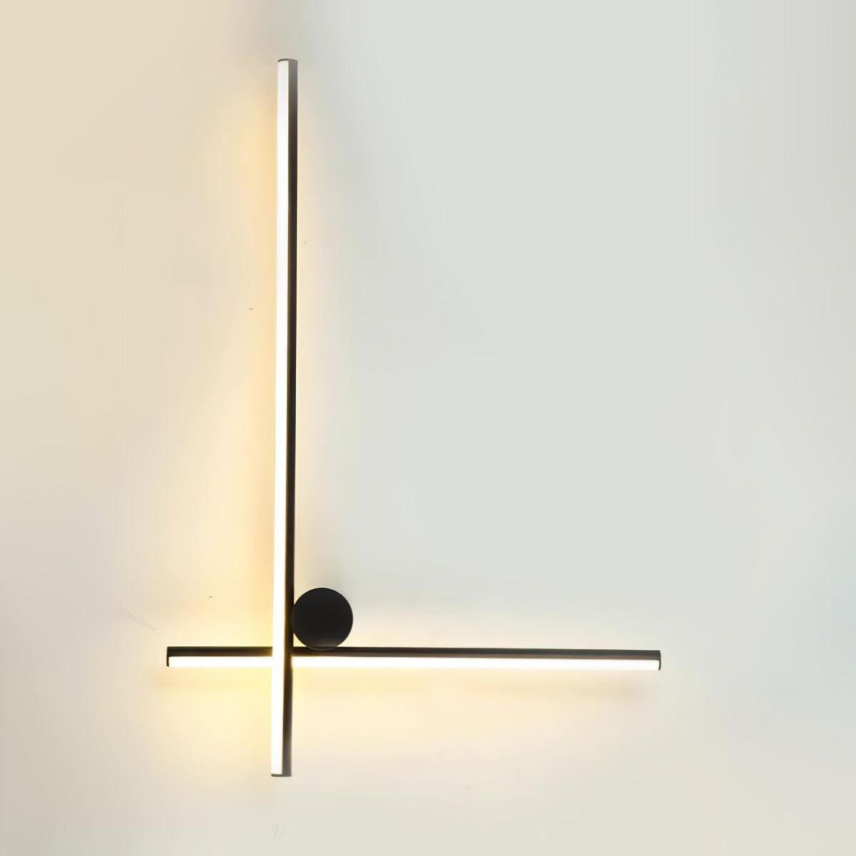 Black Clérachic wall light with a long vertical tube crossed low by a horizontal tube and a black disc, lit warm on a pale wall.