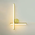 Gold Clérachic wall light with a tall vertical tube, a short horizontal tube and a large round disc, lit on a pale wall.