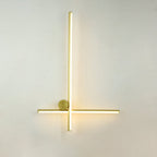 Slim gold Clérachic wall light with a tall vertical bar, a thin horizontal bar and a small round disc, lit on a pale wall.