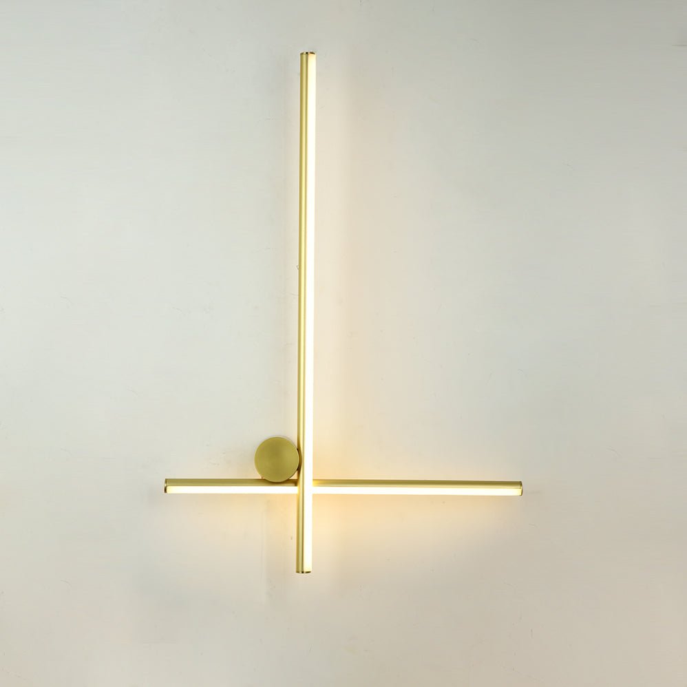 Slim gold Clérachic wall light with a tall vertical bar, a thin horizontal bar and a small round disc, lit on a pale wall.