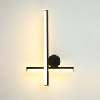 Black Clérachic wall light with a vertical tube crossed by a horizontal tube and a large black disc, lit warm on a pale wall.