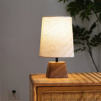 Lit Clérachic table lamp on a rattan-fronted wooden cabinet, with a leafy plant behind it and a warm glow through the linen shade.