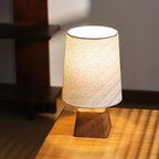 Close-ish view of this Clérachic table lamp glowing on a sunlit wooden surface, the shade's rim bright against a dark shelf.