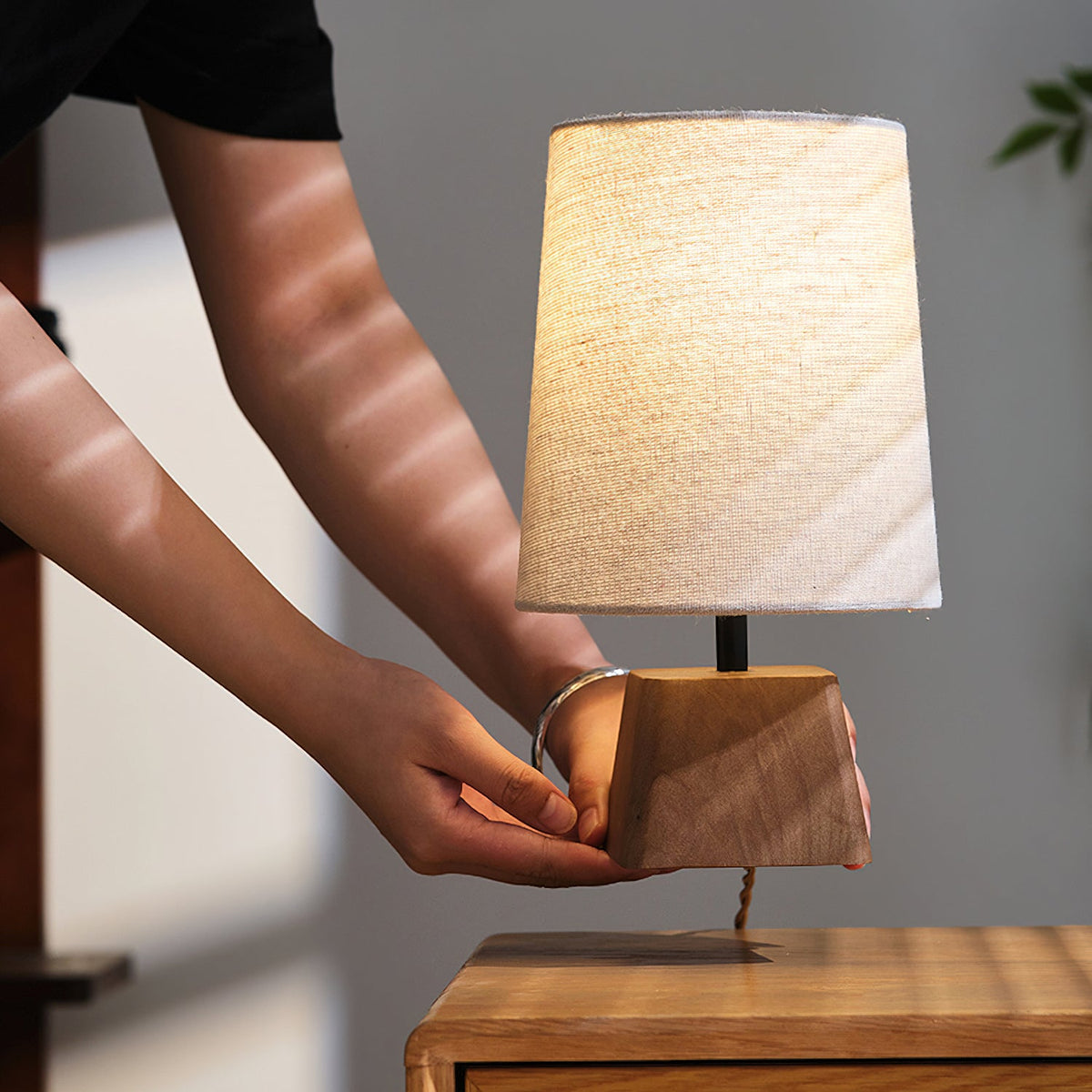 A hand steadies the wooden base of this Clérachic table lamp on a cabinet, its linen shade lit warm and soft.