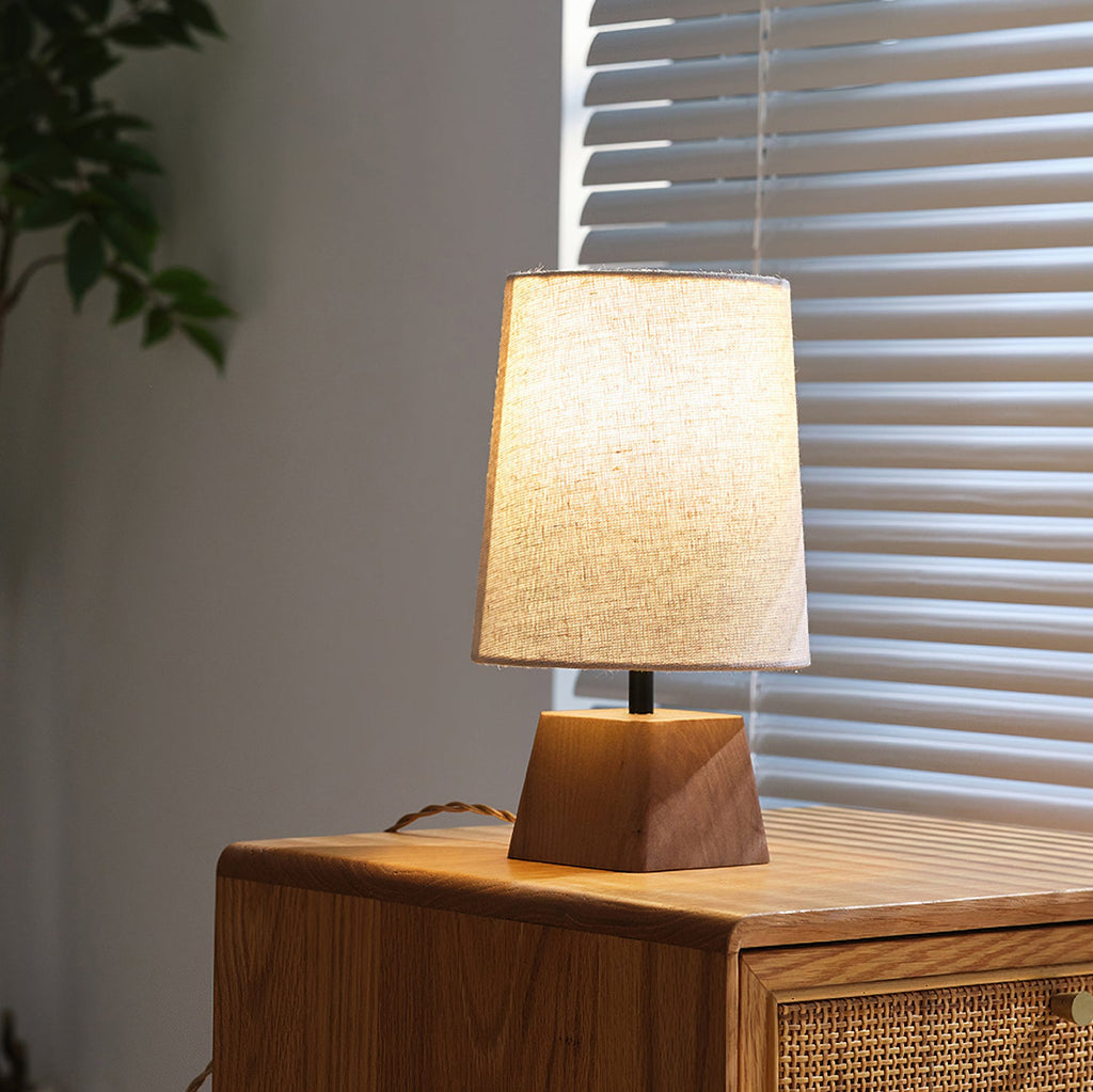 This Clérachic table lamp beside a window with slatted blinds, light glowing through its linen shade above a wooden base.