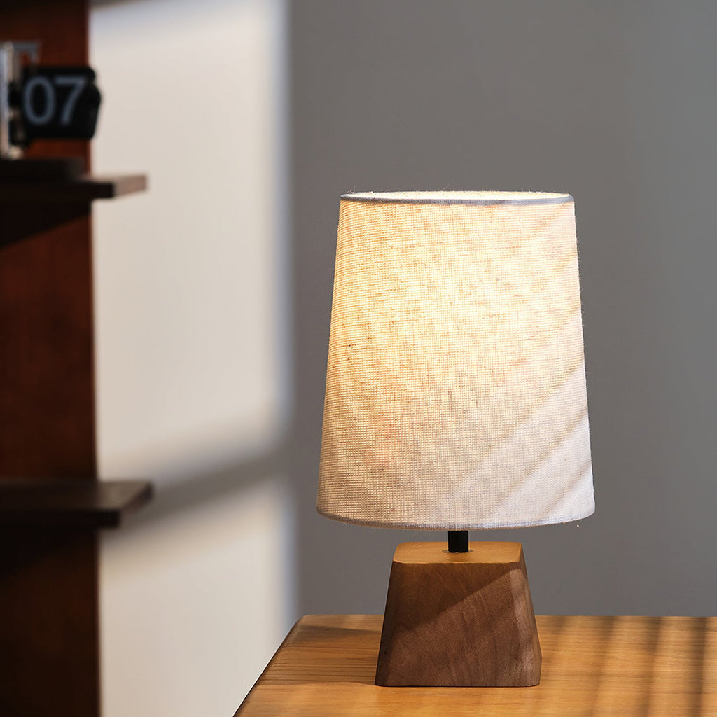 Front view of this Clérachic table lamp on a wooden surface, its tapered linen shade lit from within in a quiet grey room.