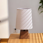 Unlit linen shade of this Clérachic table lamp, striped with diagonal sunlight, above a dark walnut-toned wooden base.