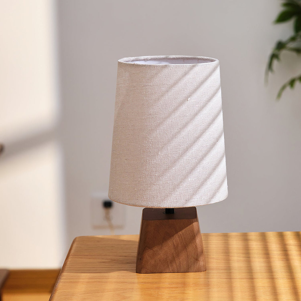 Unlit linen shade of this Clérachic table lamp, striped with diagonal sunlight, above a dark walnut-toned wooden base.