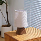 This Clérachic table lamp switched off on a wooden cabinet, a potted plant and window blinds behind its white shade.