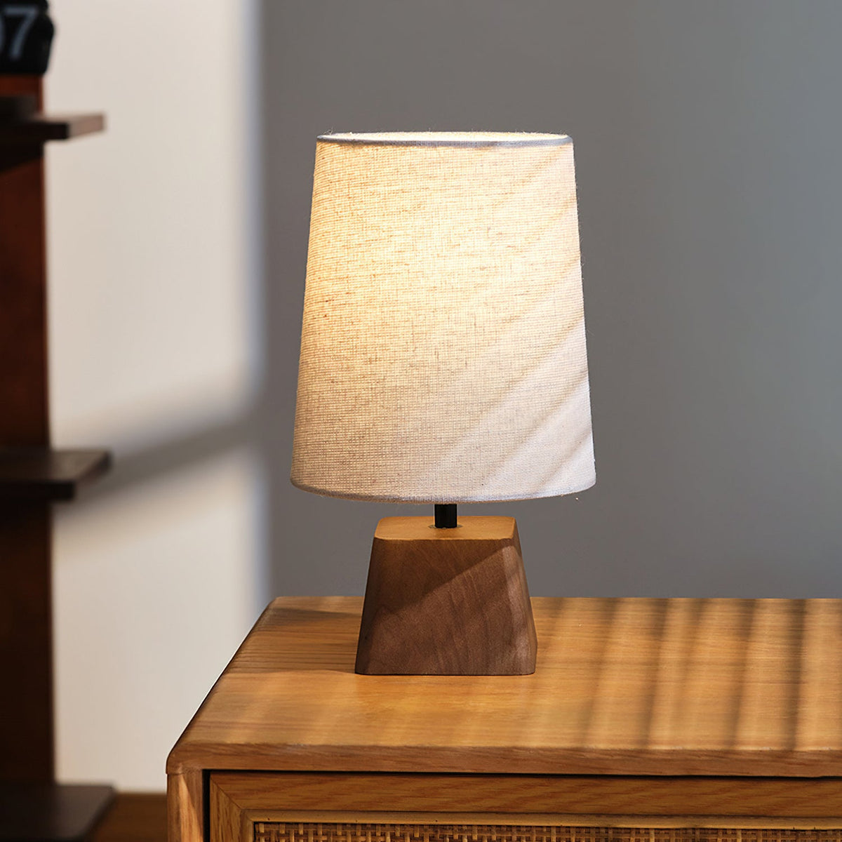Lit Clérachic table lamp on a cabinet in soft daylight, the linen shade glowing amber over its angular wooden base.
