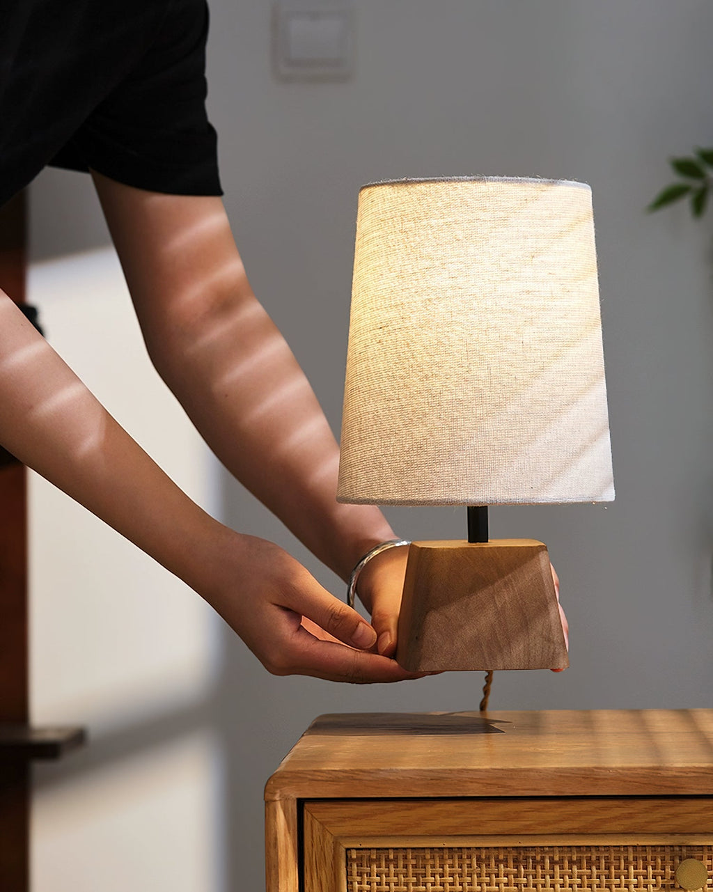 Hands set the cord beside the base of this Clérachic table lamp on a rattan-fronted cabinet, the shade softly lit.