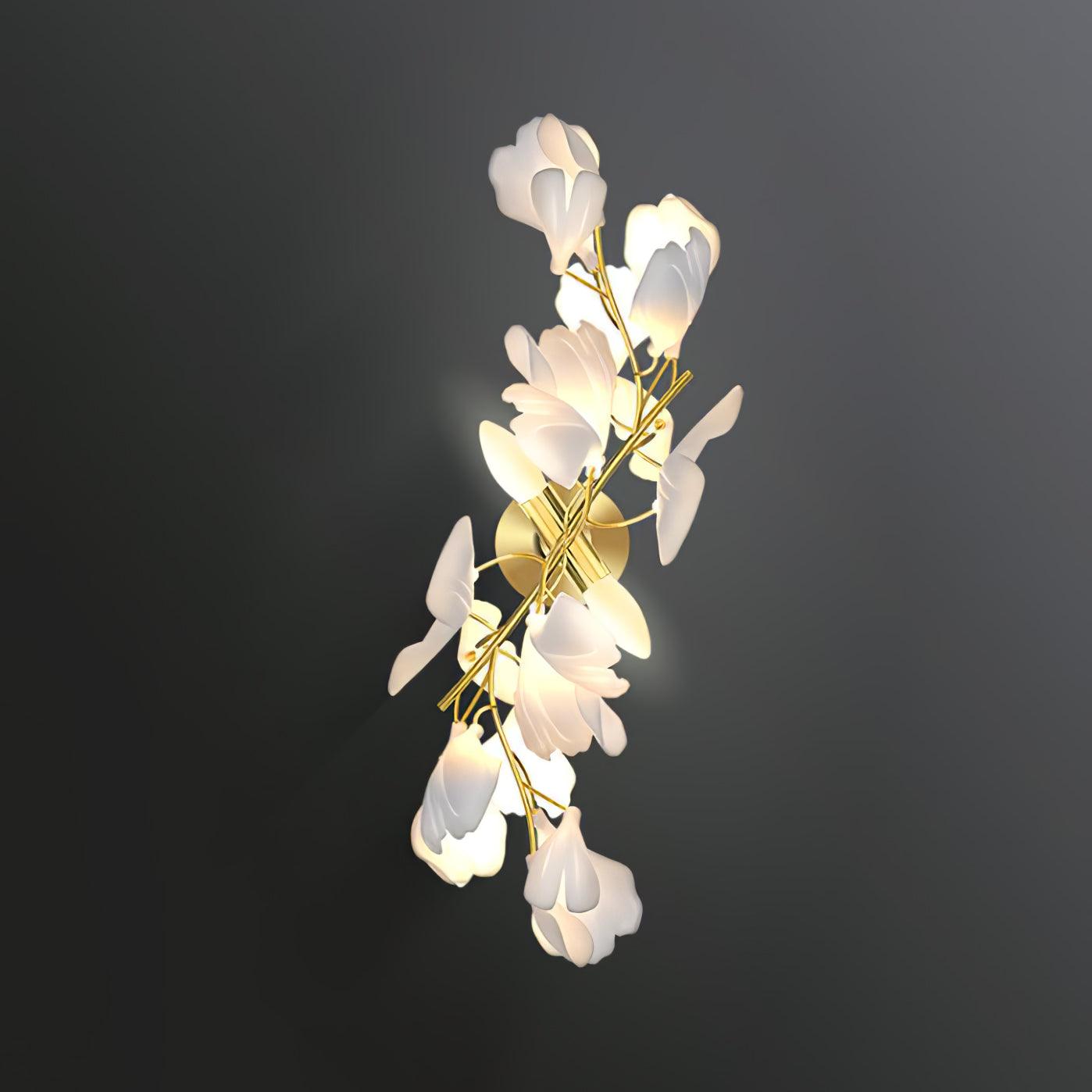 Clérachic gingko wall sconce lit on a dark wall, white ceramic petals climbing a slim gold branch.