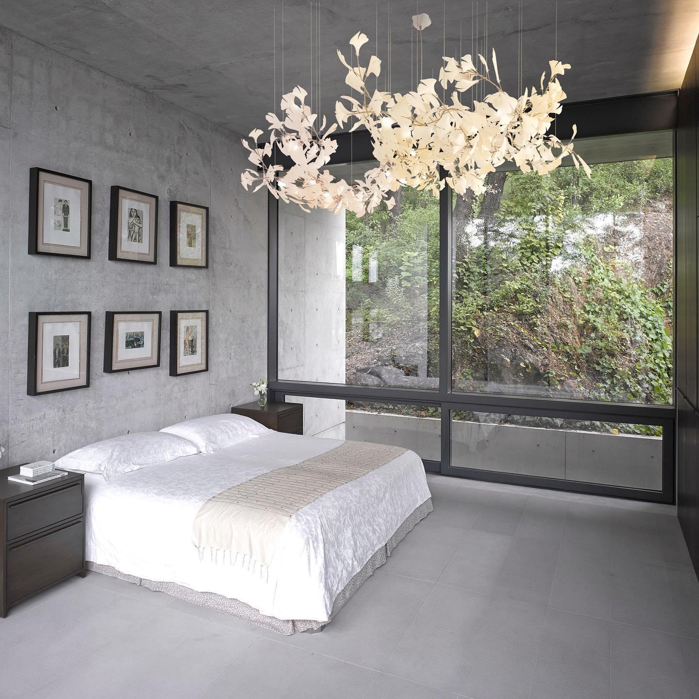 The Clérachic gingko linear chandelier lit above a bed in a concrete room with a forest view.