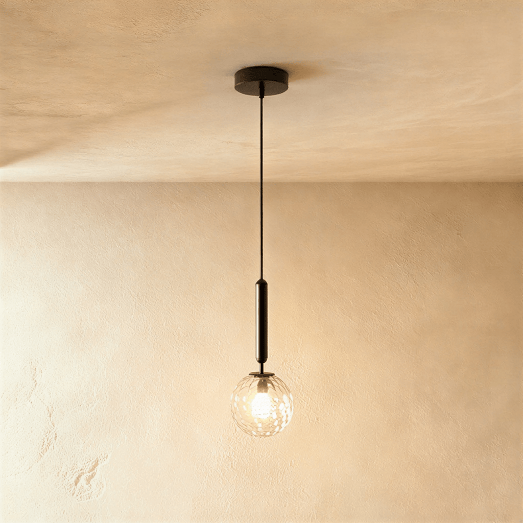 Clérachic single pendant with a clear textured glass globe and a lit bulb, on a black rod, against a warm plaster wall.