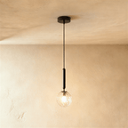 Clérachic single pendant with a clear textured glass globe and a lit bulb, on a black rod, against a warm plaster wall.