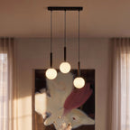 Clérachic linear pendant with three milk white glass globes on black rods, glowing before a painting and sheer curtains.