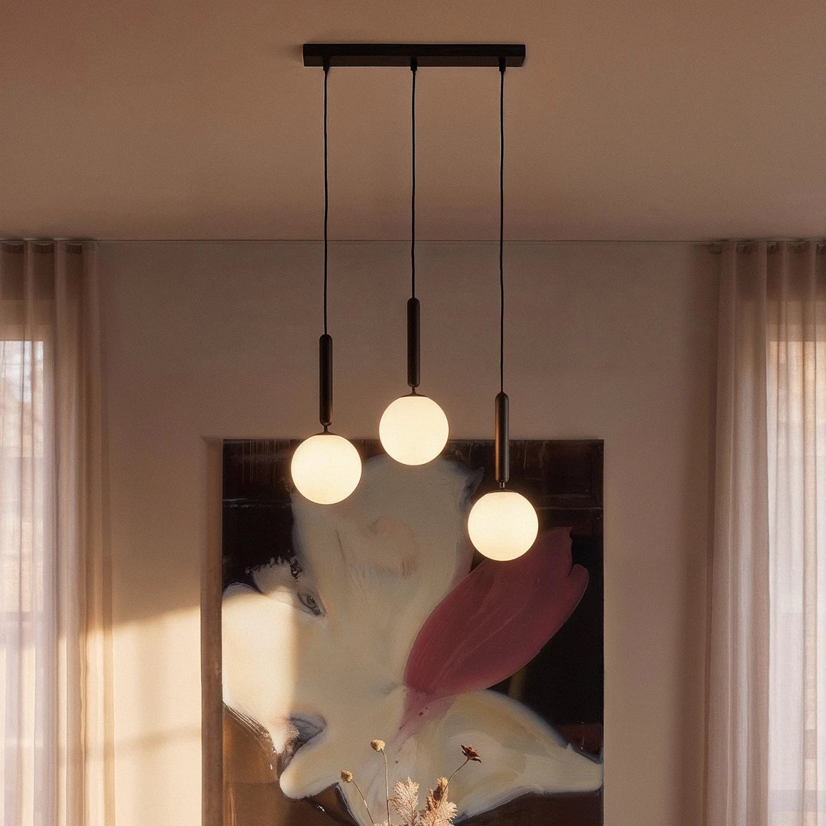 Clérachic linear pendant with three milk white glass globes on black rods, glowing before a painting and sheer curtains.