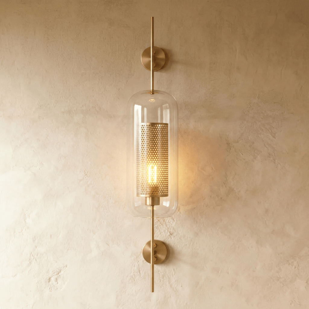 Clérachic wall lamp in the capsule shade, its perforated brass cylinder glowing behind clear glass on a warm plaster wall.