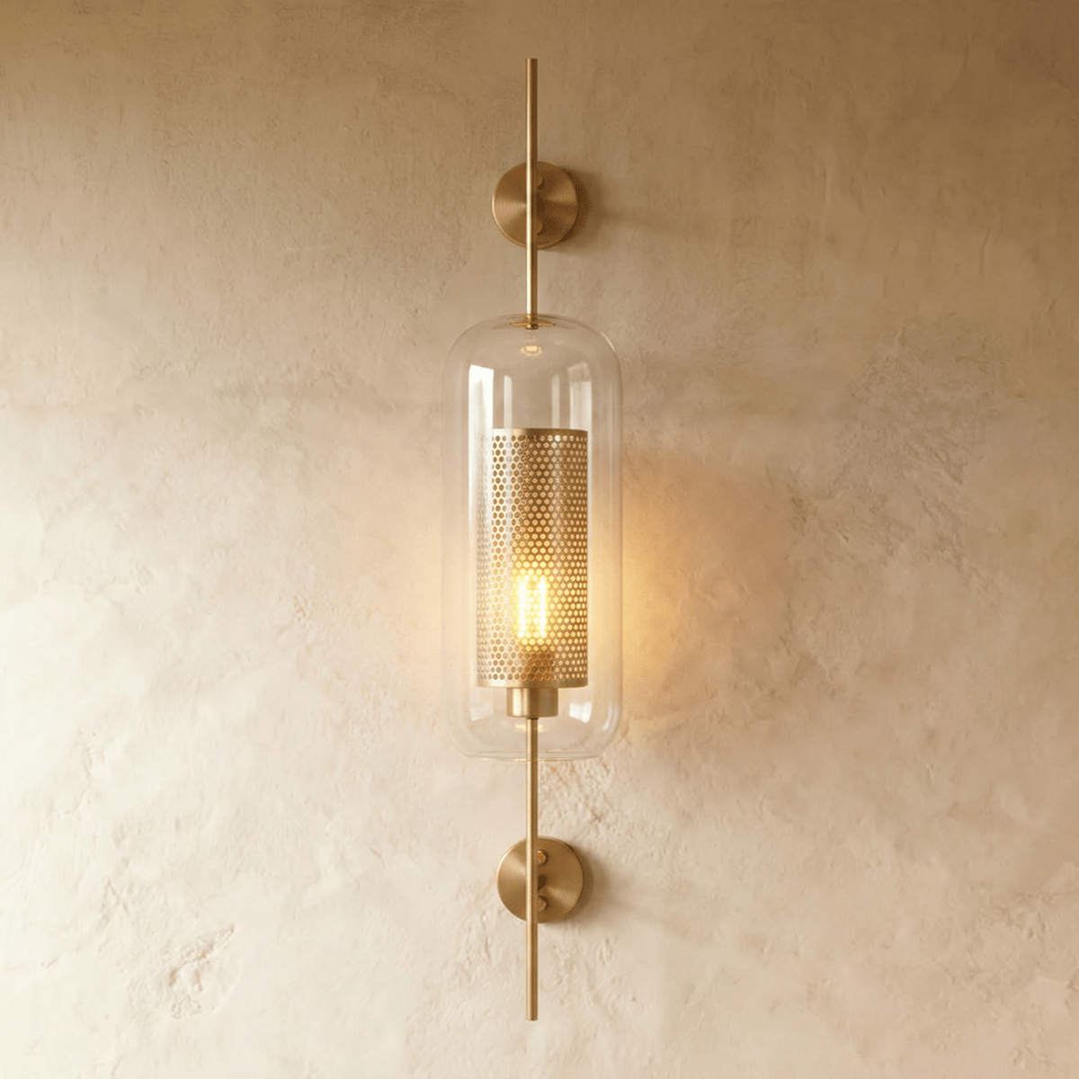 Clérachic wall lamp in the capsule shade, its perforated brass cylinder glowing behind clear glass on a warm plaster wall.