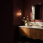 Clérachic wall lamp in the capsule shade, lit beside a marble vanity in a deep burgundy bathroom, scattering small bright dots across the wall to its left.