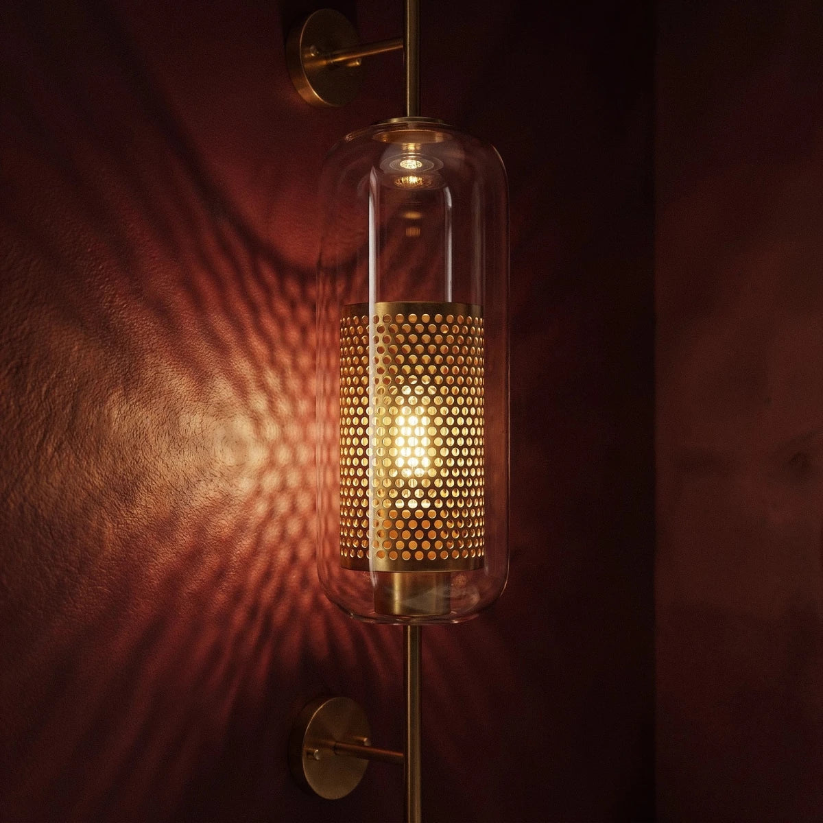 Clérachic wall lamp mounted on a textured burgundy wall, its perforated cylinder throwing a sunburst of light and shadow across the surface.