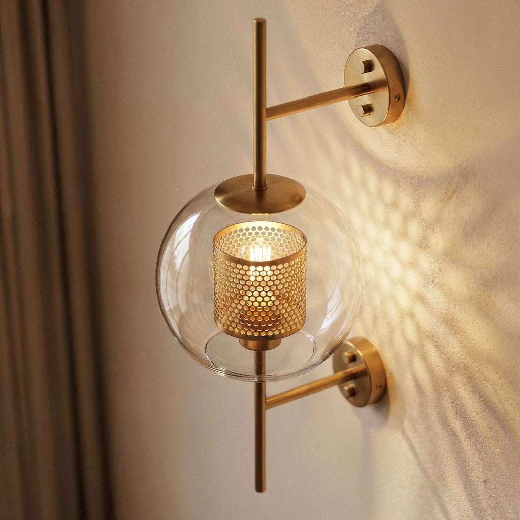Clérachic wall lamp's globe shade seen close up from a three-quarter angle, its brass cylinder punched with round holes and lit from within behind the clear glass.