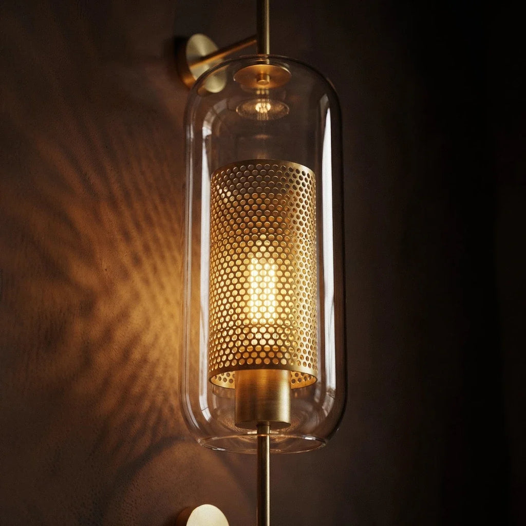 Clérachic wall lamp's capsule shade in close-up, showing the glowing bulb inside the perforated brass cylinder and the brass cap above it.