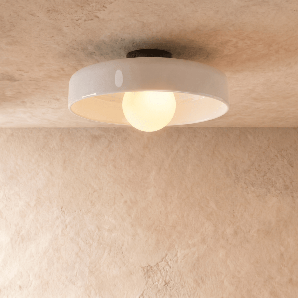 Clérachic larger white glass disc ceiling light with a glowing opal globe bulb, seen from below against a plaster wall.