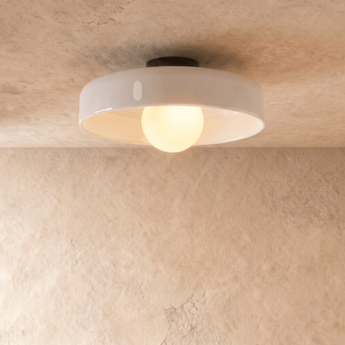 Clérachic larger white glass disc ceiling light with a glowing opal globe bulb, seen from below against a plaster wall.