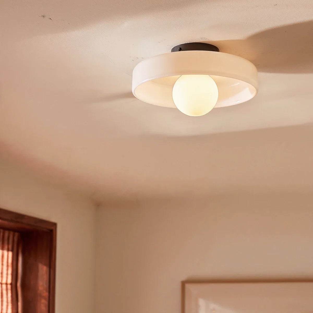Clérachic white glass disc ceiling light with an opal globe bulb, glowing warmly on a sunlit ceiling near a window.