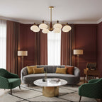 A Clérachic chandelier with white glass shades hangs in a living room with brick-red curtains, a grey curved sofa and a round marble table.
