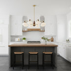 White glass Clérachic chandelier above a kitchen island with a wood top and three rattan stools, in a white shaker-style kitchen.