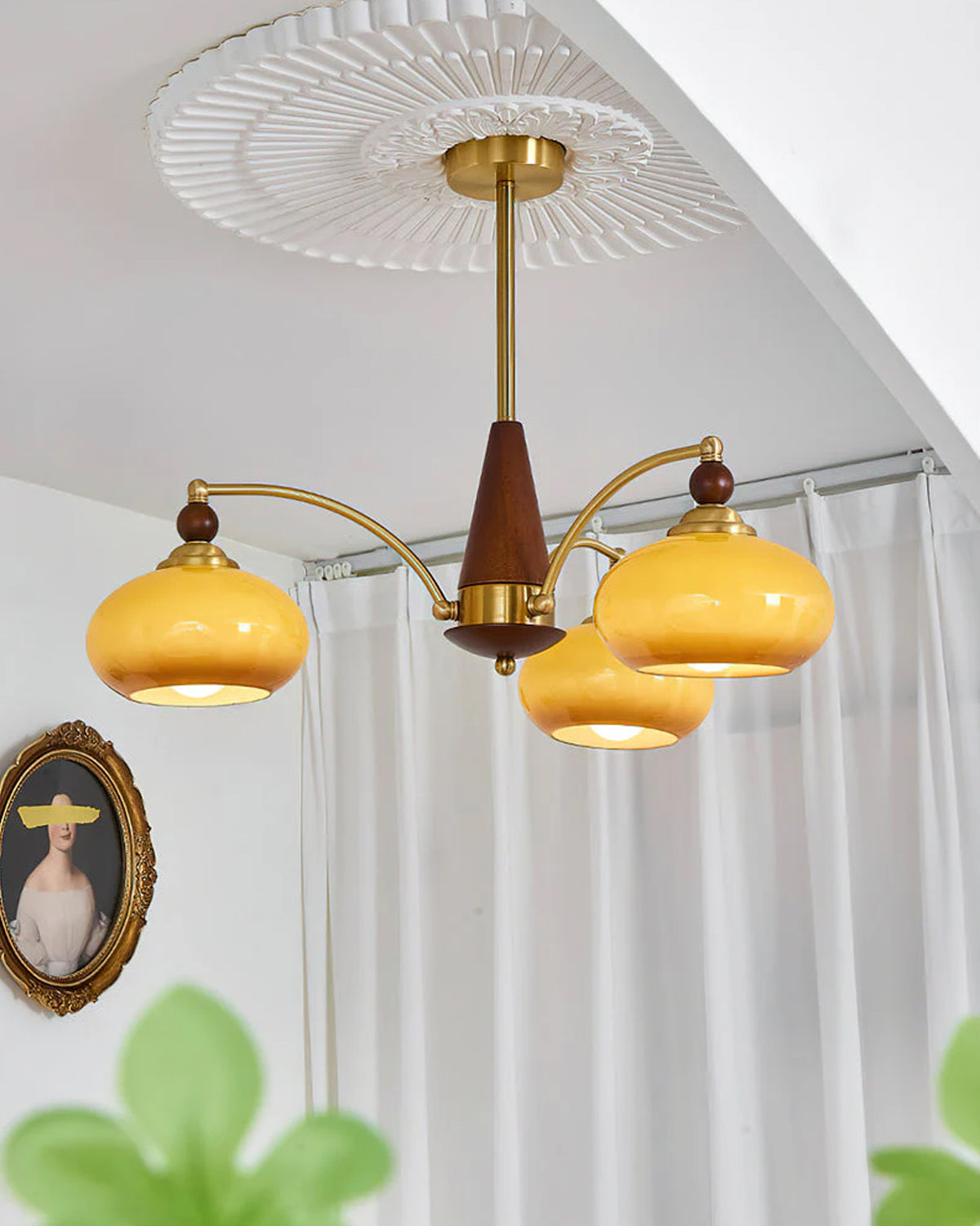 Close view of a three-head Clérachic chandelier with caramel glass shades, below a ceiling rose and in front of white curtains.