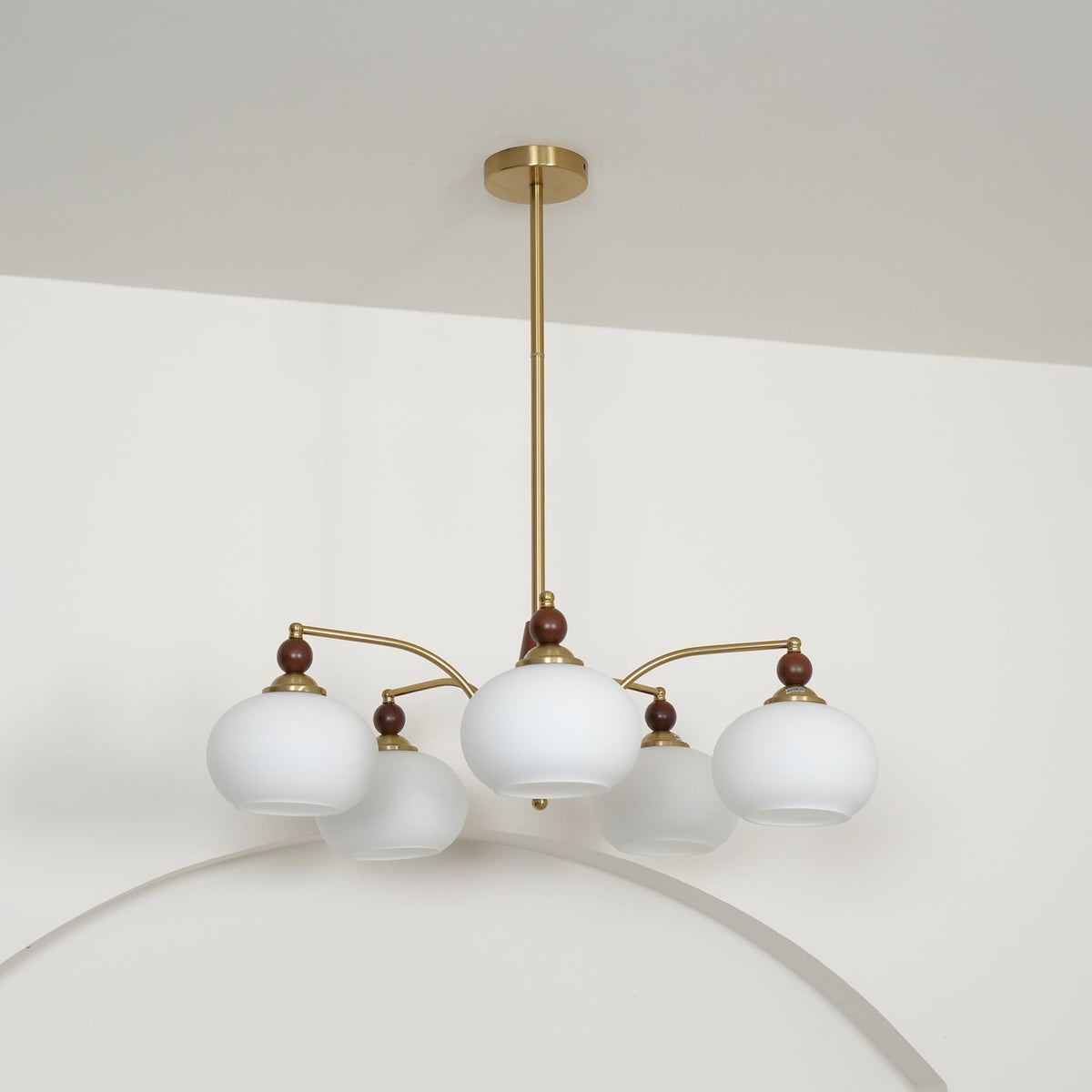 Five white glass shades, unlit, on this Clérachic chandelier, hanging from a gold ceiling plate and rod in front of a white arch.
