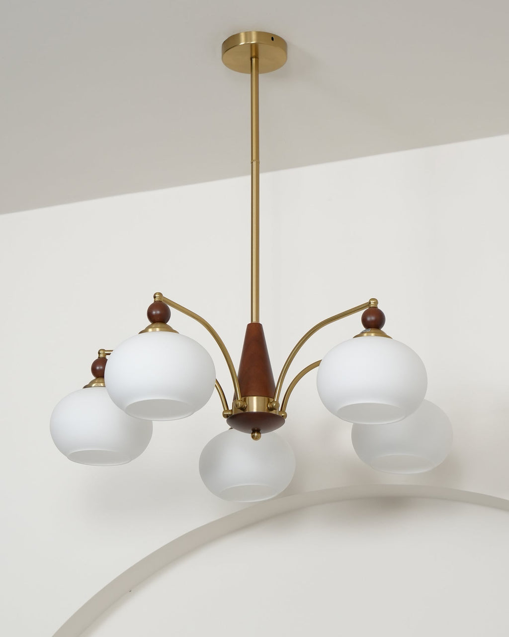 Unlit Clérachic chandelier with white glass shades, hanging from a gold rod with a ceiling plate against a white wall.