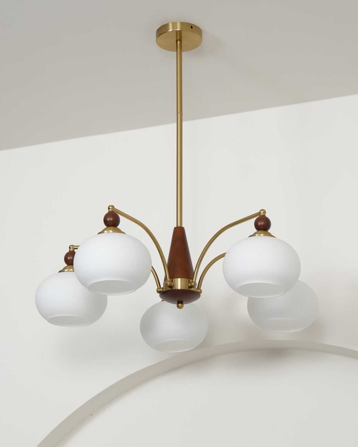 Unlit Clérachic chandelier with white glass shades, hanging from a gold rod with a ceiling plate against a white wall.